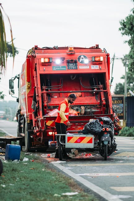 A waste collection worker wearing an orange high-visibility vest and black trousers is seen operating a large red rubbish truck on a street, positioned with its rear open for loading waste. The truck, belonging to Waste Clearance Edgware, is parked partly on the pavement and partly on the roadway, with visible safety markings on the vehicle's back. In front of the truck, the worker is handling a black plastic garbage bag, which is resting on a small cart or platform attached to a motorbike that is parked nearby. The scene is set in an urban environment with a green grassy area and trees visible in the background, along with a clear sky and some overhead power lines. The street is relatively quiet, with some distant signage and buildings visible, indicating a typical street scene where private waste collection and rubbish removal services are being carried out. The overall setting emphasizes on-site rubbish clearance, aligning with the context of professional waste management services such as those provided by Waste Clearance Edgware.