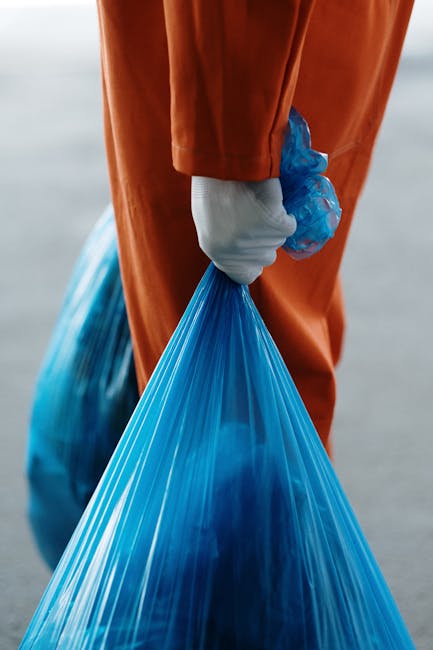 A person wearing an orange waterproof suit and white gloves is holding a tightly knotted blue plastic rubbish bag by its gathered top handle. The bag appears full and is made of opaque, glossy plastic, with its contents creating a rounded bulge. In the background, part of a similar blue bag is visible, partially resting on the ground or pavement. The environment looks like an outdoor area, possibly a street or a designated waste collection point, with a neutral, slightly blurred backdrop that emphasizes the individual and the bags. The scene reflects a typical rubbish collection or waste disposal activity, highlighting the process of on-site waste handling or preparation for private rubbish removal services such as those provided by Waste Clearance Edgware. The image underscores the importance of secure bagging for safe and efficient rubbish removal, aligning with professional waste management practices, especially for those opting for independent collection outside local authority services.