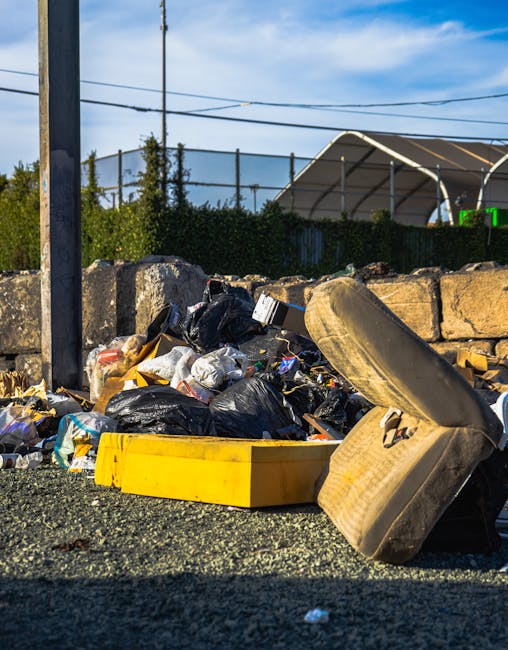 A pile of mixed household waste and rubbish situated on a paved surface in an outdoor area, including black plastic rubbish bags, a yellow plastic container, and a discarded old beige fabric armchair with visible dirt and wear. The debris is piled against a low stone wall made of irregularly shaped rocks, with some waste spilling onto the ground, indicating an informal or private rubbish collection scene. In the background, there is a wire fence with green foliage, and further behind, a sports court with a curved roof structure and some overhead power lines are visible under a partly cloudy sky. The scene reflects an instance of on-site waste clearance, possibly conducted by a private waste removal service, with the environment suggesting a suburban or urban setting ready for rubbish collection or disposal. Waste Clearance Edgware is familiar with handling such bulk refuse. Natural lighting highlights the textures and colors of the waste materials, emphasizing a casual yet organized approach to rubbish clearance, suitable for customers seeking alternative waste handling methods outside of standard municipal services.