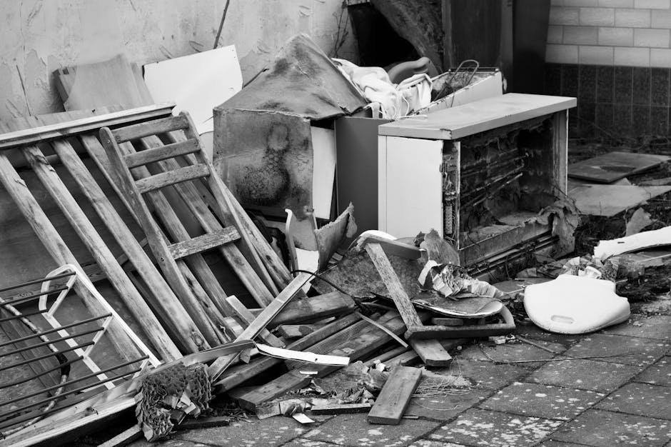 A pile of discarded rubbish and debris is shown outside a building, consisting of wooden furniture, including a tilted wooden bed frame with multiple slats, which appears weathered and worn, with visible grain and aged texture. Adjacent to the frame is a white appliance, possibly an old refrigerator or storage unit, with its door slightly ajar, revealing a dark interior. Resting on or around the appliance are various broken materials such as torn cardboard, damaged wood, and scattered small debris. A white plastic object, likely a part of household waste like a basin or container, is lying on the ground near the rubble. The background shows a brick wall and concrete pavement, with the scene illuminated by natural daylight. The environment suggests an outdoor area where private waste clearance or on-site rubbish disposal has occurred, possibly facilitated by Waste Clearance Edgware as part of a bulky rubbish removal or alternative waste handling service.