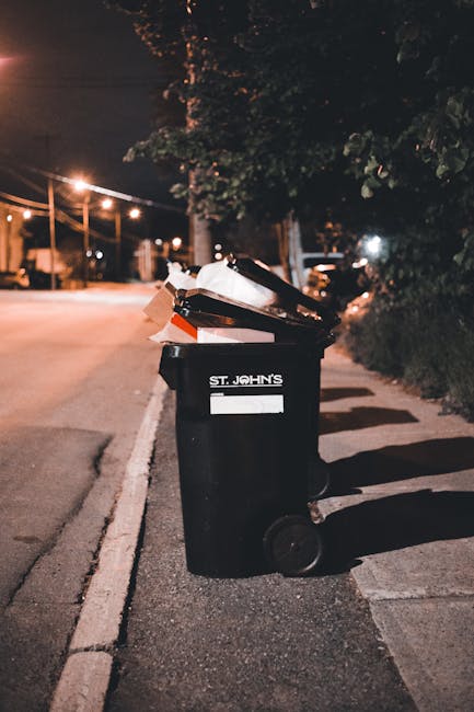 A black wheelie bin labeled 'St. John's' is positioned on a quiet residential street at night, standing on the pavement near the curb. The bin is filled with various waste items, including paper, cardboard, and plastic, some of which are overflowing and slightly protruding from the open lid. The street is illuminated by warm streetlights, casting soft light and shadows on the bin and surrounding pavement. In the background, a row of trees partly obscures a hedge, with faint outlines of houses and parked vehicles visible further along the street. The scene suggests a typical late evening period, with the bin possibly awaiting collection through an independent rubbish removal or local waste disposal service, such as Waste Clearance Edgware. The environment appears calm with no pedestrians, and the overall setting highlights the practical aspect of residential rubbish storage before scheduled collection or alternative waste handling actions.