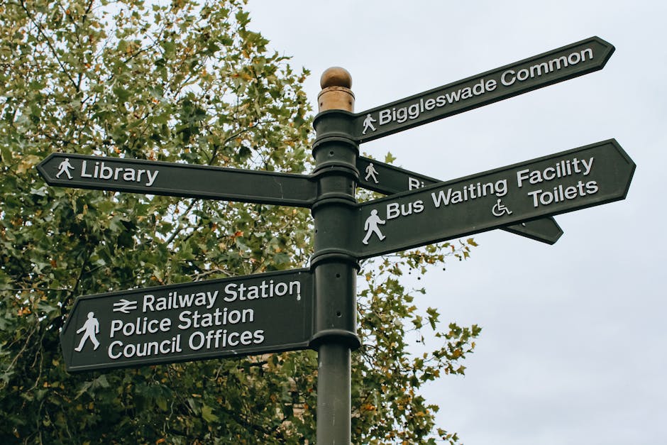 A black directional signpost with multiple rectangular signs pointing in different directions, mounted on a dark pole. The signs display white text indicating nearby locations including 'Biggleswade Common,' 'Library,' 'Bus Waiting Facility,' 'Toilets,' 'Railway Station,' 'Police Station,' and 'Council Offices.' Some signs feature icons such as pedestrians and wheelchair accessibility symbols. The background features a large leafy tree with green foliage, partially obscuring a light grey sky. The scene appears to be outdoors in a public area, possibly within or near a park or town centre. The signpost's clean, modern design contrasts with the natural surroundings, representing a typical arrangement used for local wayfinding and navigation, contributing to the context of effective signage in public spaces.