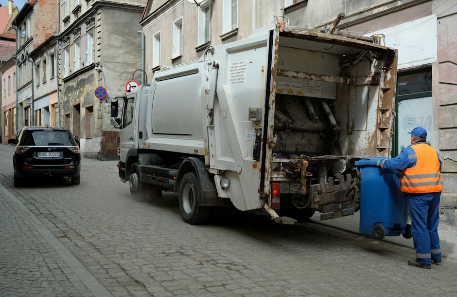 A waste collection truck with a white body and a rusted, open rear compartment is parked on a cobblestone street in an urban area. The truck's rear loading section contains no visible rubbish but shows signs of wear and rust. A worker wearing a blue uniform, orange high-visibility vest, and blue protective gloves is positioned to the right of the truck, emptying a blue wheeled bin into the vehicle. The street is lined with multi-storey residential buildings displaying a mix of stone and plaster facades, with windows and some peeling paint. In the background, a parked black car is visible along with urban signage, including a no-parking sign. The scene is lit with natural daylight, and the overall environment suggests a typical municipal rubbish collection operation, which can be part of on-site clearance or private waste handling services, as managed by waste clearance providers like Waste Clearance Edgware.