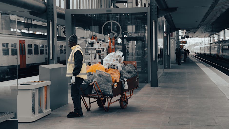 A waste collection truck with a white body and a rusted, open rear compartment is parked on a cobblestone street in an urban area. The truck's rear loading section contains no visible rubbish but shows signs of wear and rust. A worker wearing a blue uniform, orange high-visibility vest, and blue protective gloves is positioned to the right of the truck, emptying a blue wheeled bin into the vehicle. The street is lined with multi-storey residential buildings displaying a mix of stone and plaster facades, with windows and some peeling paint. In the background, a parked black car is visible along with urban signage, including a no-parking sign. The scene is lit with natural daylight, and the overall environment suggests a typical municipal rubbish collection operation, which can be part of on-site clearance or private waste handling services, as managed by waste clearance providers like Waste Clearance Edgware.