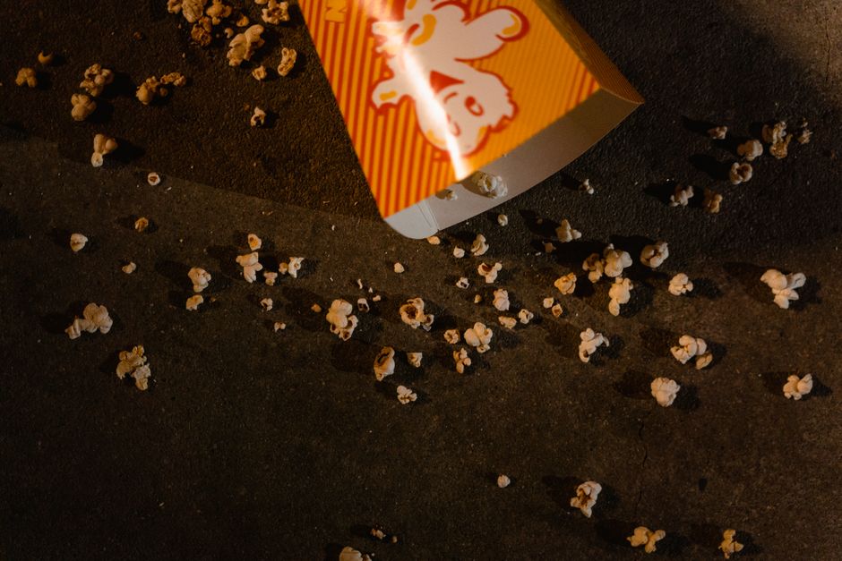 A close-up image shows a partially visible, tilted cardboard popcorn container with a red and yellow striped design, featuring an illustrated clown face. The container is placed on a dark, rough-textured surface, likely asphalt or concrete, illuminated by artificial lighting that creates shadows and highlights. Small popped popcorn pieces are scattered across the surface, some near the container and others spread out, with irregular shapes and a light, creamy color. The scattered popcorn and container suggest a scene of discarded snack packaging, possibly after a public event or outdoor gathering, with no other objects or background elements visible in the frame. This image may relate to waste management or rubbish removal services, illustrating the need for on-site clearance of food waste and litter, such as those provided by Waste Clearance Edgware, on urban pavements or public spaces.