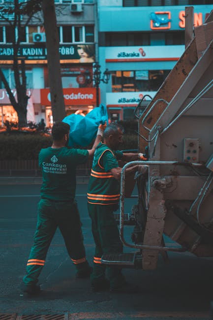 Two waste collection workers dressed in dark green uniforms with reflective orange stripes are positioned beside a large industrial waste collection vehicle on an urban street. One worker, on the left, stands with his back to the camera and is lifting a bright blue plastic bag filled with rubbish, preparing to load it into the vehicle. The other worker, on the right, faces the vehicle and appears to be operating or overseeing the loading process. The waste vehicle, situated on the right side of the image, features a metallic, painted surface with hydraulic components visible, and is parked on a paved street. In the background, there is a busy commercial area with illuminated signs, including telecommunications and clothing stores, and storefronts with large glass windows. The street is lined with leafless trees, suggesting it might be autumn or winter, and the ambient lighting indicates early evening or dusk. Waste Clearance Edgware’s team is conducting on-site rubbish removal in an urban environment, supporting private waste disposal services that complement or offer alternatives to local authority collection, with waste being deliberately loaded into the vehicle for disposal or transfer.