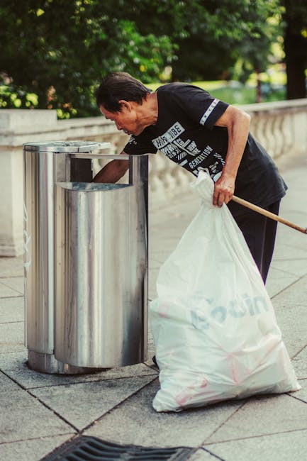A large plastic waste receptacle with a blue lid and a white label marked 'mixed paper & card' is overflowing with various rubbish items, including scattered cardboard boxes, plastic bags, paper waste, and packaging materials, some of which are spilling onto the surrounding pavement. To the left of the bin, there are additional black and red rubbish bags, while several flattened cardboard boxes and assorted discarded items lie on the ground nearby. Behind the waste containers, a parked grey car with yellow license plates is visible, with a metal railing separating the parking area from the pavement. In the background, a modern building with scaffolding, blue paneling, and glass windows is present, alongside signs indicating commercial premises, including a fish bar. The scene is outdoors on a cloudy day, with the waste accumulation highlighting a contrast to regular rubbish collection services and illustrating a situation that could be associated with private or on-site waste clearance providers like Waste Clearance Edgware.